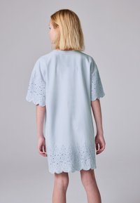 Svetle modré šaty s krátkými, vlnitými rukávy a složitou výšivkou s průstřihy podél lemu, zobrazené zezadu.