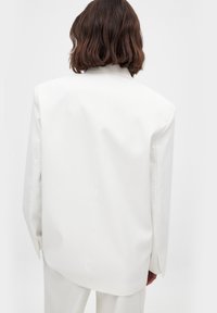 Blazer bianco con design su misura, spalle strutturate e una superficie liscia. La vista posteriore mostra un orlo leggermente sceso e una cucitura minimalista.