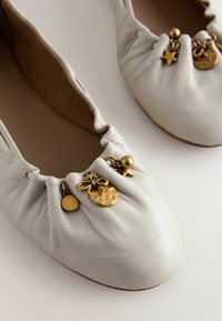 Balto odinio baleto bateliai su surinktų raukšlių detalėmis prie pirštų, papuošti auks tono pakabukais: žvaigžde, kamuoliuku ir tekstūruota skrituliu.