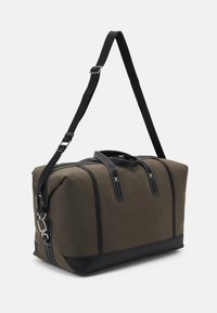 Sac de voyage en toile couleur khaki avec des accents en cuir noir, poignées doubles et bandoulière réglable. Il présente une couture blanche contrastante et une fermeture éclair.