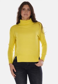 Maglione giallo a collo alto realizzato in tessuto morbido, con una vestibilità comoda, polsini e orlo a costine. Presenta spacchi laterali per un maggiore comfort. Indossato con jeans scuri.