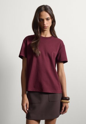 Joven mujer con cabello largo y castaño, que lleva una camisa de manga corta de color rojo oscuro y una falda marrón oscuro, de pie frente a un fondo claro y liso.