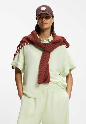 Kobieta w jasnoszmaragdowej koszuli z krótkim rękawem i spodniach, z rdzawej barwy dzianinowym swetrem przewieszonym przez ramiona oraz ciemnobrązową czapką skierowaną do przodu.