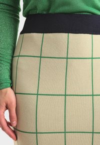 Béžová tužková sukně s zeleným kostkovaným vzorem, která má tmavě námořnický elastický pas. Tkanina má hladký, strukturovaný vzhled.