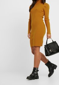 Hořčicový šaty s ribovaným vzorem, dlouhými rukávy a vysokým límcem; doplněné černými kotníkovými botami se sponami a černou texturovanou kabelkou.