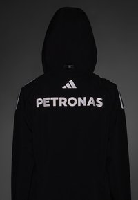 Sort hættejakke med sølv "PETRONAS" logo og Adidas emblem på ryggen, med en glat tekstur og minimalistisk design.