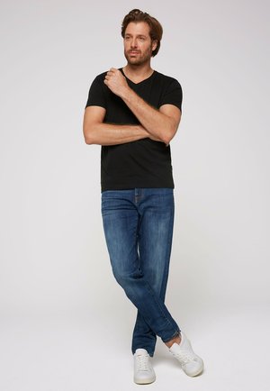 Black short-sleeve V-neck t-shirt paired with faded blue jeans and white sneakers, standing with arms crossed against a plain background.