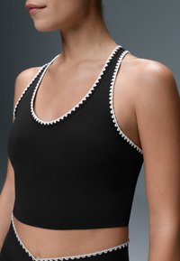 Woman wearing a black sleeveless crop top with scalloped white trim along the neckline and armholes, shown from shoulders to waist.