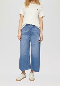 Vaaleanbeige t-paita, jossa on pieni musta ja sininen sydänkuvio, yhdistettynä leveälahkeisiin, vaaleansinisiin farkkuihin ja valkoisiin lenkkareihin.