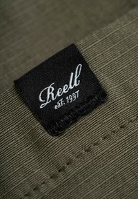 Tejido verde oliva con una etiqueta negra cosida, que presenta el texto cursivo "Reell" y "est. 1997." Superficie texturizada con costuras visibles.