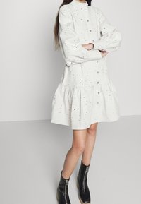 Robe chemise en jean blanc avec une jupe à volants, dotée d'un col montant, de boutons-pression et de détails en œillets. Portée avec des bottines noires.
