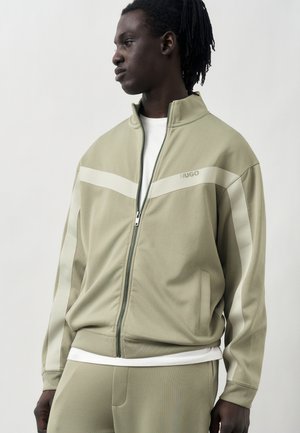 Mann trägt eine beige Reißverschlussjacke und passende Hose mit weißen Streifendetails, blickt nach links vor einfachem Hintergrund.