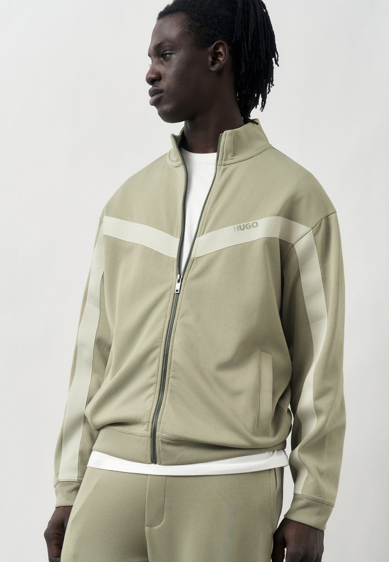 Άνδρας που φοράει μπεζ μπουφάν με φερμουάρ και ταιριαστό παντελόνι με λευκές ρίγες, κοιτάζει αριστερά σε απλό φόντο.