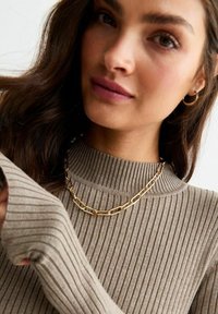 Maglione beige a costine con colletto alto, che mette in mostra una collana in oro e orecchini a cerchio. Morbida texture e vestibilità aderente dettagliata nel tessuto.