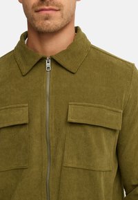 Olivengrønt kordfløjljakke med lynlås foran, spidse kraver og to brystlommer. Stoffet har en ribbet tekstur.