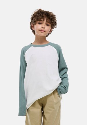 Garçon aux cheveux bouclés portant un t-shirt à manches longues blanc et vert ainsi qu'un pantalon beige, debout avec une main dans la poche, sur un fond uni.