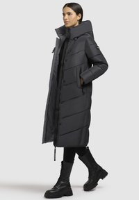 Lang sort pufferjakke med høj krave, quiltet mønster og forlommer. Båret med sorte leggings og kampstøvler.