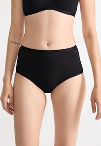 Femme portant des sous-vêtements noirs taille haute et un soutien-gorge noir, montrant le torse et la partie supérieure des cuisses devant un fond blanc uni.