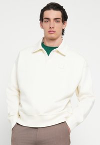 Felpa bianca con scollo a polo e polsini a coste. Presenta un logo discreto al centro ed è abbinata a una maglietta verde sottostante.