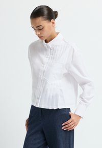 Camicia bianca con bottoni, caratterizzata da un design plissettato su un lato, taschino sul petto e colletto strutturato. Abbinata a pantaloni scuri a righe sottili.