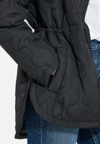 Chaqueta negra acolchada con una cintura ajustada, bolsillos laterales y una textura de tela suave. Presentada con jeans azules.
