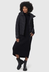 Black puffer jacket with hood, quilted design, worn over a black ribbed knit dress, paired with black chunky-soled ankle boots.