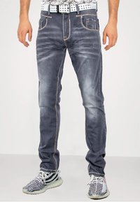 Dunkelgraue Denim-Jeans mit leichter Abnutzung, kontrastierendem orangefarbenem Stitching, Fünf-Taschen-Design und schmaler Passform. Getragen mit gemusterten Sneakers.