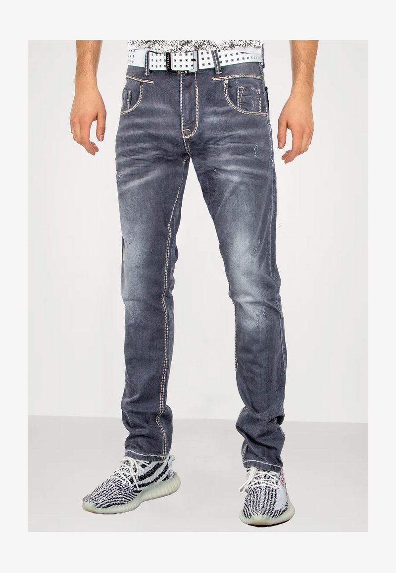 Dunkelgraue Denim-Jeans mit leichter Abnutzung, kontrastierendem orangefarbenem Stitching, Fünf-Taschen-Design und schmaler Passform. Getragen mit gemusterten Sneakers.