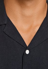 Camicia navy a maniche corte con collo, caratterizzata da un singolo bottone bianco e un tessuto testurizzato. Vista ravvicinata dell'area del colletto.