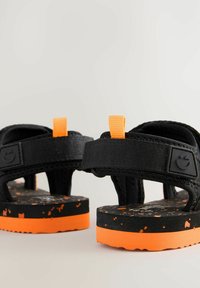 Mustat sandaalit oransseilla yksityiskohdilla, joissa on teksturoitu hihna, säädettävä takaosa ja pilkullinen musta-oranssi pohja.