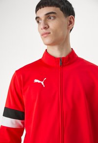 Rød idrettsjakke laget av glatt stoff, med høy krage, glidelås og en hvit Puma-logo på venstre bryst. Svart og hvite striper på ermene.