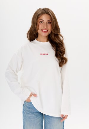 Witte blouse met lange mouwen en een ronde hals, voorzien van rode geborduurde tekst "MIRONS" op de borst. G gedragen met lichtblauwe jeans.