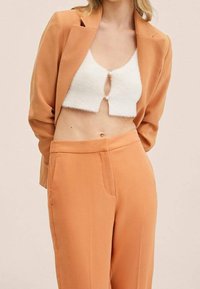 Orange blazer over en hvid cropped top med detalje af frontknapper, parret med matchende orange bukser. Glat stof med skræddersyet pasform.