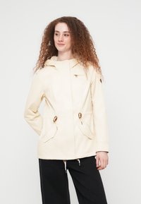 Femme aux cheveux roux bouclés portant une veste beige à capuche avec des poches avant et un pantalon noir, regardant légèrement vers sa droite.