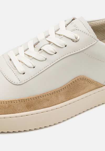 Nærbillede af en cremefarvet lædersneaker med hvide snørebånd og en lys brun ruskindsdetalje langs siden nær sålen.