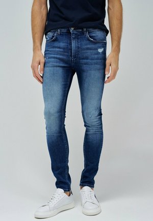 Vaqueros slim-fit azules con un ligero desgaste y detalles desgastados. Hechos de denim, con bolsillos clásicos y cierre de cremallera. Llevados con zapatillas blancas.