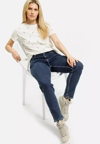 Biała koszulka z krótkim rękawem z małym kwiatowym nadrukiem, zestawiona z ciemnoniebieskimi dżinsami. Model siedzi na krześle, nosząc jasnobeżowe sneakersy.