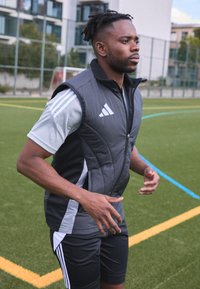 Un individuo atlético con un chaleco acolchado negro y gris con cremallera, una camiseta gris de mangas cortas y pantalones cortos negros, en un campo de césped verde.