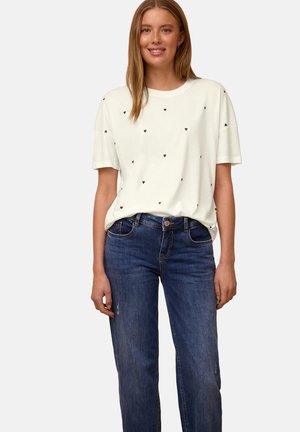 Vrouw draagt een wit T-shirt met korte mouwen met kleine zwarte hartjes en een blauwe jeans, staand tegen een effen lichte achtergrond.