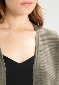 Olive zielenie sweter rozpinany z prążkowanym wzorem, z otwartym przodem i miękkiego materiału, noszony na czarnej bluzce z dekoltem w serek.