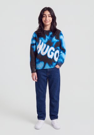 Ung person iført en blå og sort sweater med teksten ""HUGO"", mørkeblå jeans og hvide sneakers, med den ene hånd i lommen.