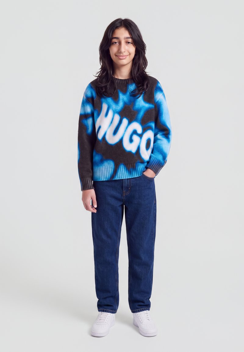 Jeune personne portant un pull bleu et noir avec le texte ""HUGO"", un jean bleu foncé et des baskets blanches, une main dans la poche.