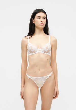 Hosszú sötét hajú nő fehér, virágos csipke melltartóban és hozzáillő bugyiban, egyszerű világos háttér előtt.