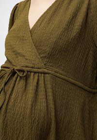 Gros plan d'une personne portant une robe portefeuille texturée vert olive avec un décolleté en V et une ceinture nouée à la taille.