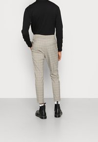 Person som bär beige rutiga byxor, svart långärmad tröja, vita strumpor och svarta ankelboots som står mot en neutral vägg.