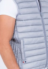 Gilet rembourré gris clair avec des coutures verticales, fermeture éclair et poches sur les côtés. Présente une étiquette de marque en rouge, blanc et bleu sur l'ourlet.