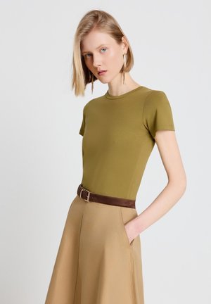 Donna con capelli corti biondi che indossa una maglietta aderente verde oliva a maniche corte, gonna beige a vita alta, cintura marrone e lunghi orecchini pendenti.
