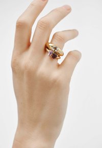 Anillos de oro en una mano: un aro liso con una piedra clara, el otro presenta piedras preciosas rosas y marrones, mostrando formas y colores variados.