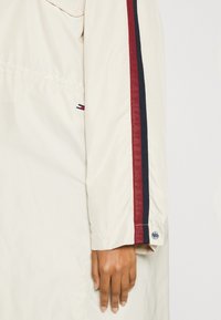 Beige lehká bunda s červenými a námořnickými pruhy na rukávech, která má knoflíkové zapínání na manžetách a jemný detail loga.