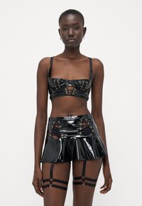 Čierny latexový crop top so zlatými očkami a výrezmi, doplnený čiernou plisovanou sukňou s korzetovým šnurovaním a traky.